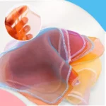 Une pile d'écharpes vibrantes et translucides aux bords cousus de blanc provenant de la Boîte à Mouchoirs Magique ; un encart montre une main étirant une écharpe pour en montrer la texture.