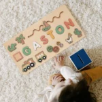 Un enfant interagit avec un Puzzle Personnalisé Prénom en Bois - Un souvenir unique comportant le nom « GASTON » et diverses pièces de bois colorées, dont un train, des cactus et des formes d'animaux, sur un tapis beige.