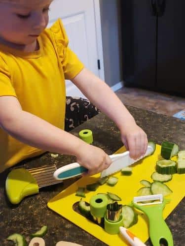 Un jeune enfant en chemise jaune coupe des concombres sur une planche à découper jaune à l'aide d'un emporte-pièce ondulé Couteaux Enfant. Des ustensiles de cuisine et des morceaux de concombre sont visibles sur le comptoir.