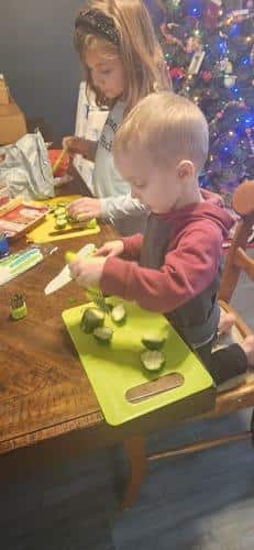 Un jeune garçon et une jeune fille utilisent des Couteaux Enfant pour couper des concombres sur des planches à découper vertes à une table, avec un sapin de Noël décoré en arrière-plan.