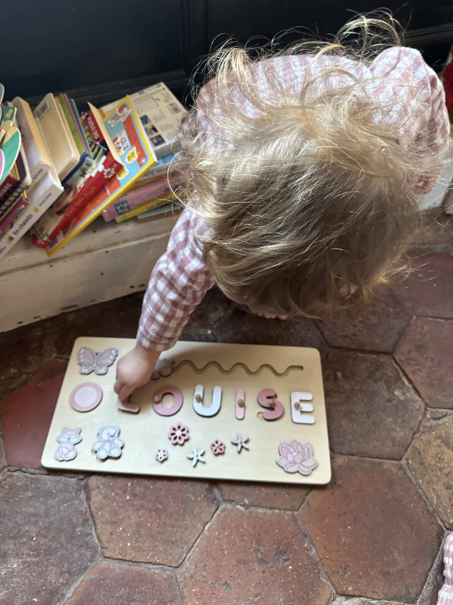 Un enfant en pyjama à carreaux roses dispose les pièces d'un puzzle en bois portant le nom « LOUISE » sur un sol carrelé près d'une pile de livres.