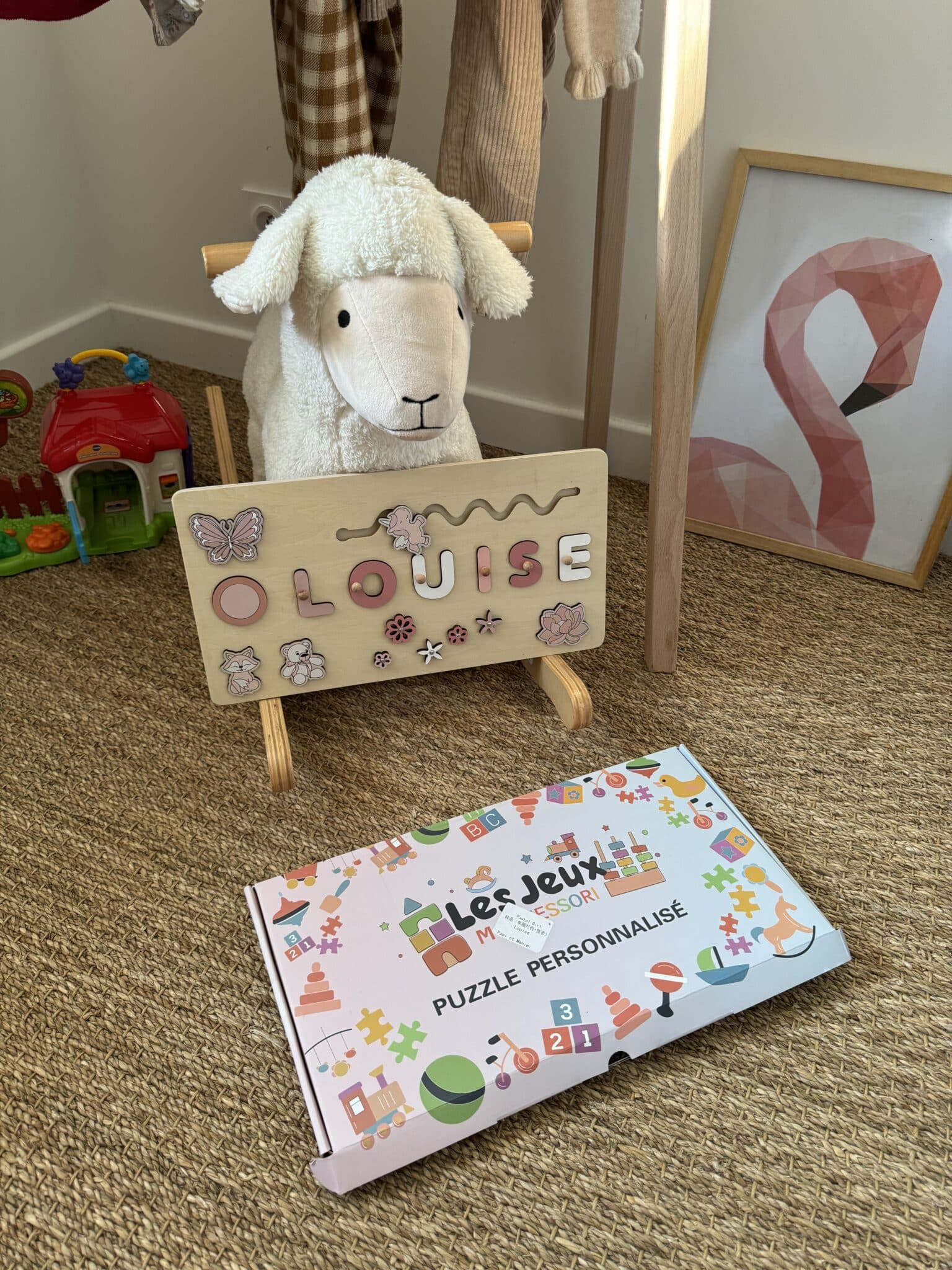Un mouton en peluche à bascule est posé derrière une pancarte en bois portant le nom « LOUISE ». Devant, une boîte colorée est étiquetée « Puzzle Personnalisé ». Des jouets et des œuvres d'art pour enfants sont à l'arrière-plan.