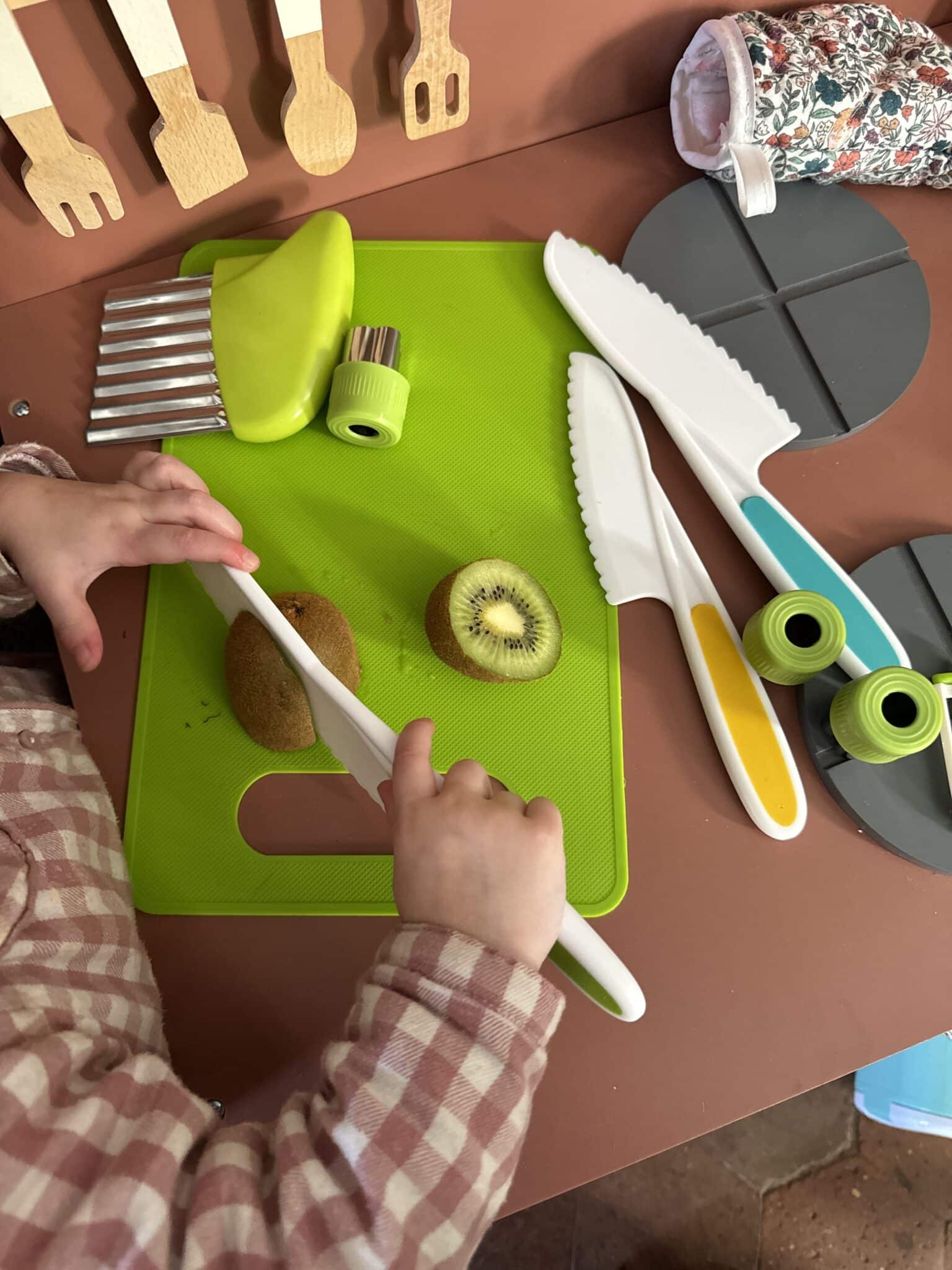Un enfant utilise un Couteaux Enfant pour couper un kiwi sur une planche à découper verte, entouré de divers ustensiles de cuisine.