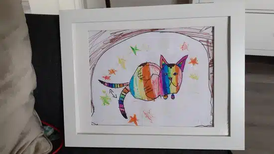 Dessin encadré (Cadre Dessin) d'un chat rond aux couleurs de l'arc-en-ciel entouré d'étoiles colorées, exposé sur un canapé sombre avec des oreillers en arrière-plan.