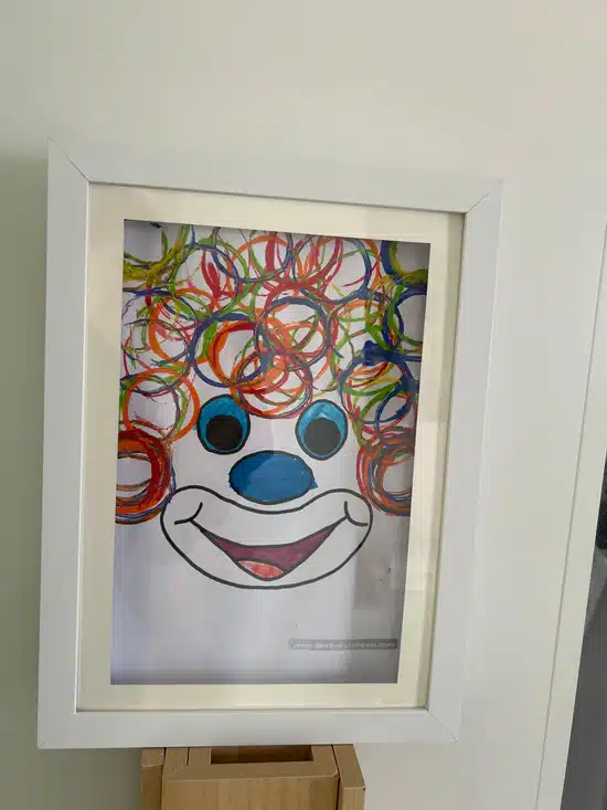 Cadre encadré Dessin d'un visage de clown avec des gribouillis colorés et circulaires pour les cheveux, un grand nez bleu, des joues bleues et une large bouche souriante.