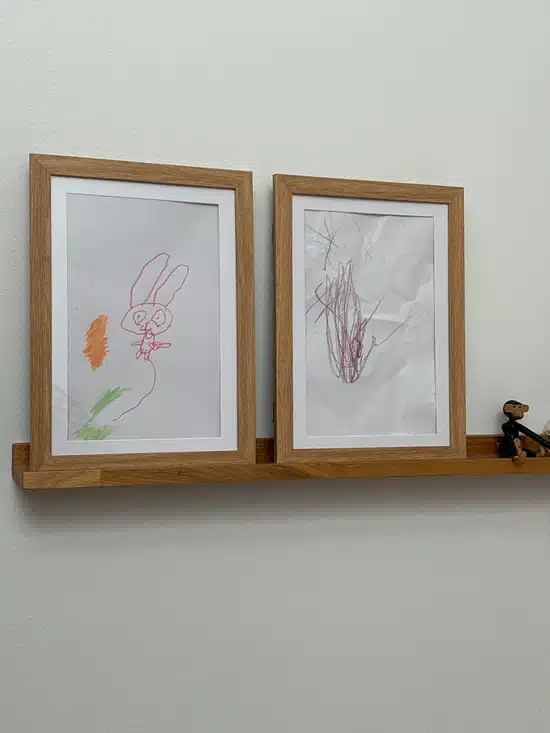 Deux dessins d'enfants encadrés sont exposés sur une étagère en bois contre un mur blanc, avec une petite figurine placée à l'extrémité droite de l'étagère.