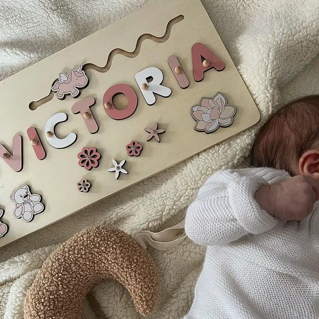 Un puzzle en bois portant le nom "VICTORIA" avec des décorations florales et animales est posé sur une couverture à côté d'un bébé vêtu de blanc et d'un jouet texturé en forme de croissant.