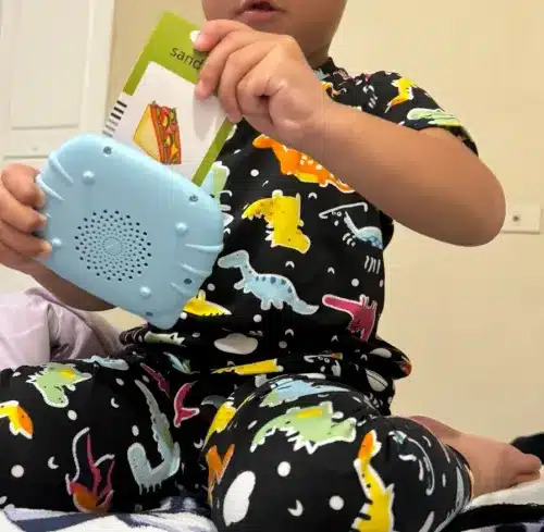 Un enfant en pyjama dinosaure utilise un appareil bleu portable, tout en serrant contre lui une carte illustrant un sandwich. Ces premiers moments sont à la fois attachants et éphémères.