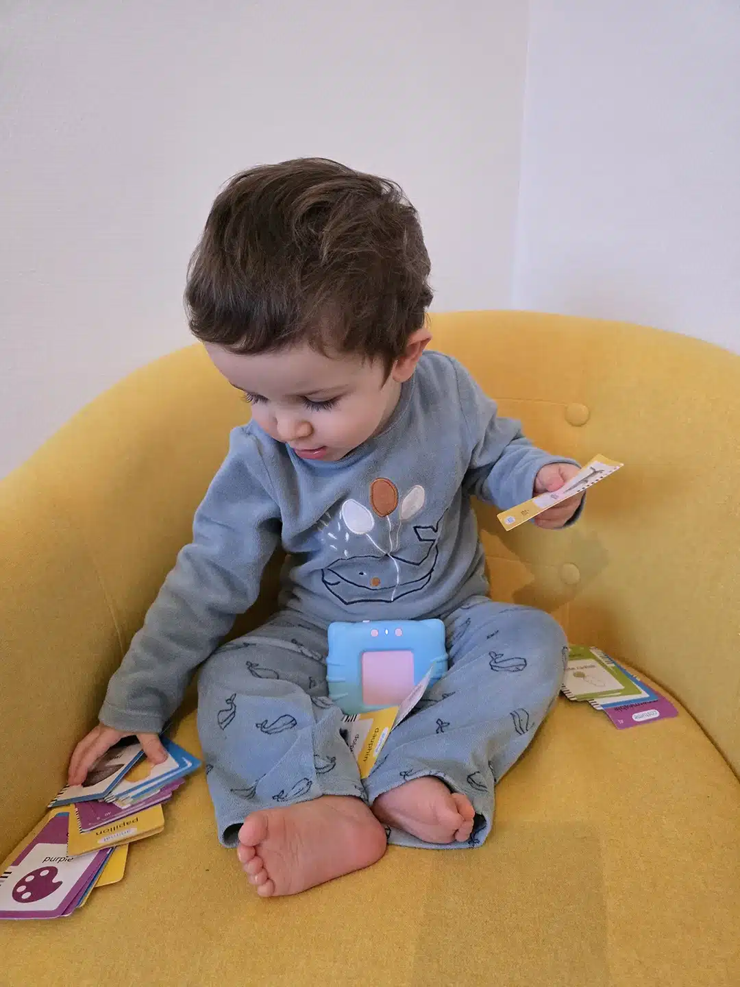 Un bambin en pyjama bleu, explorant ses premiers mots, est assis sur une chaise jaune entouré de cartes à jouer, en tenant deux en main.