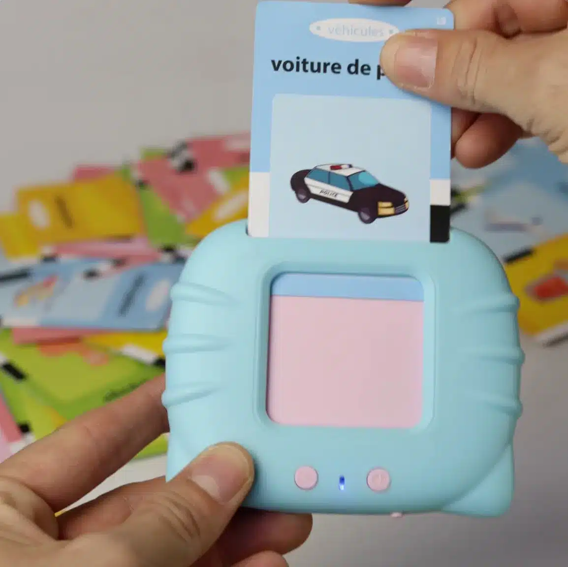 Une personne insère une carte de « voix de police » dans un dispositif pédagogique bleu de la collection Premiers Mots. D’autres cartes colorées sont dispersées en arrière-plan.