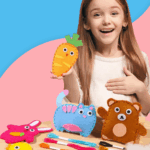 Une petite fille sourit en tenant une carotte en feutre qu'elle a fabriquée avec le Kit de Couture Enfants - Porte-clés à Faire Soi-Même en Sécurité. Des formes d'animaux en feutre, du fil et du matériel de bricolage sont disposés sur la table devant elle.