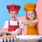 Deux enfants portent le Tablier et Toque de Cuisine pour Enfant. Ils sourient en cuisinant avec de la farine, des œufs et des ustensiles sur la table, devant un fond coloré.