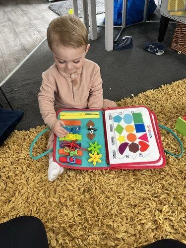 Un jeune enfant est assis sur un tapis, interagissant avec un livre éducatif coloré présentant des formes et des textures, un peu comme un kit sensoriel encourageant l'exploration tactile.