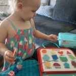 Un jeune enfant vêtu d'une robe à motifs interagit avec un puzzle de formes Kit Sensoriel sur la table, explorant des lettres et des formes vibrantes.