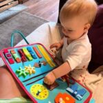 Un bébé est assis sur un canapé et explore joyeusement un tableau d'activités coloré, qui fait partie d'un kit sensoriel innovant. Ce tableau stimulant est doté de divers éléments interactifs, dont une horloge et des formes, conçus pour stimuler les jeunes esprits.