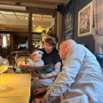 Une femme, berçant un bébé et un livre ouvert, est assise à une table, se livrant à une expérience multisensorielle. Un homme se penche pour admirer le bébé, créant une atmosphère chaleureuse et décontractée dans la salle à manger intérieure.