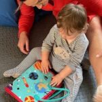 Un adulte et un enfant, assis par terre, explorent un livre coloré aux illustrations sur le thème de l'espace. Le pyjama rayé de l'enfant est assorti aux tons vibrants de son kit sensoriel, créant une expérience sensorielle captivante.