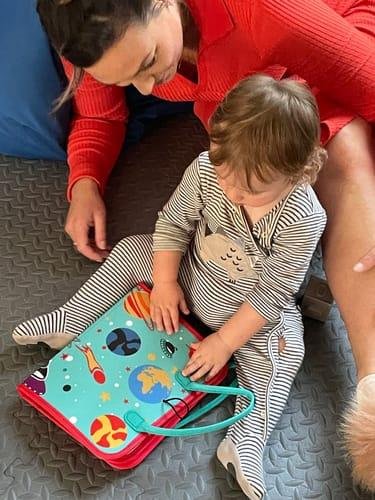 Un adulte et un enfant, assis par terre, explorent un livre coloré aux illustrations sur le thème de l'espace. Le pyjama rayé de l'enfant est assorti aux tons vibrants de son kit sensoriel, créant une expérience sensorielle captivante.