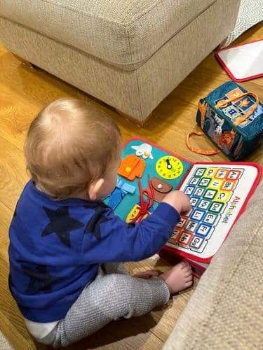 Un tout-petit vêtu d'un pull bleu est assis sur un parquet et joue avec un livre éducatif en tissu coloré de son kit sensoriel. Le livre comprend une section alphabet et divers éléments interactifs qui captivent sa curiosité.