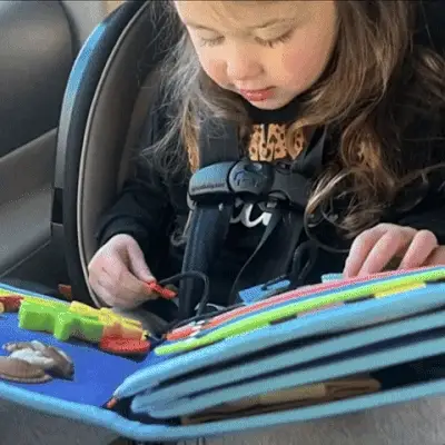 Un jeune enfant dans un siège auto est captivé par un kit sensoriel coloré, rempli de petits jouets et d'activités engageantes.