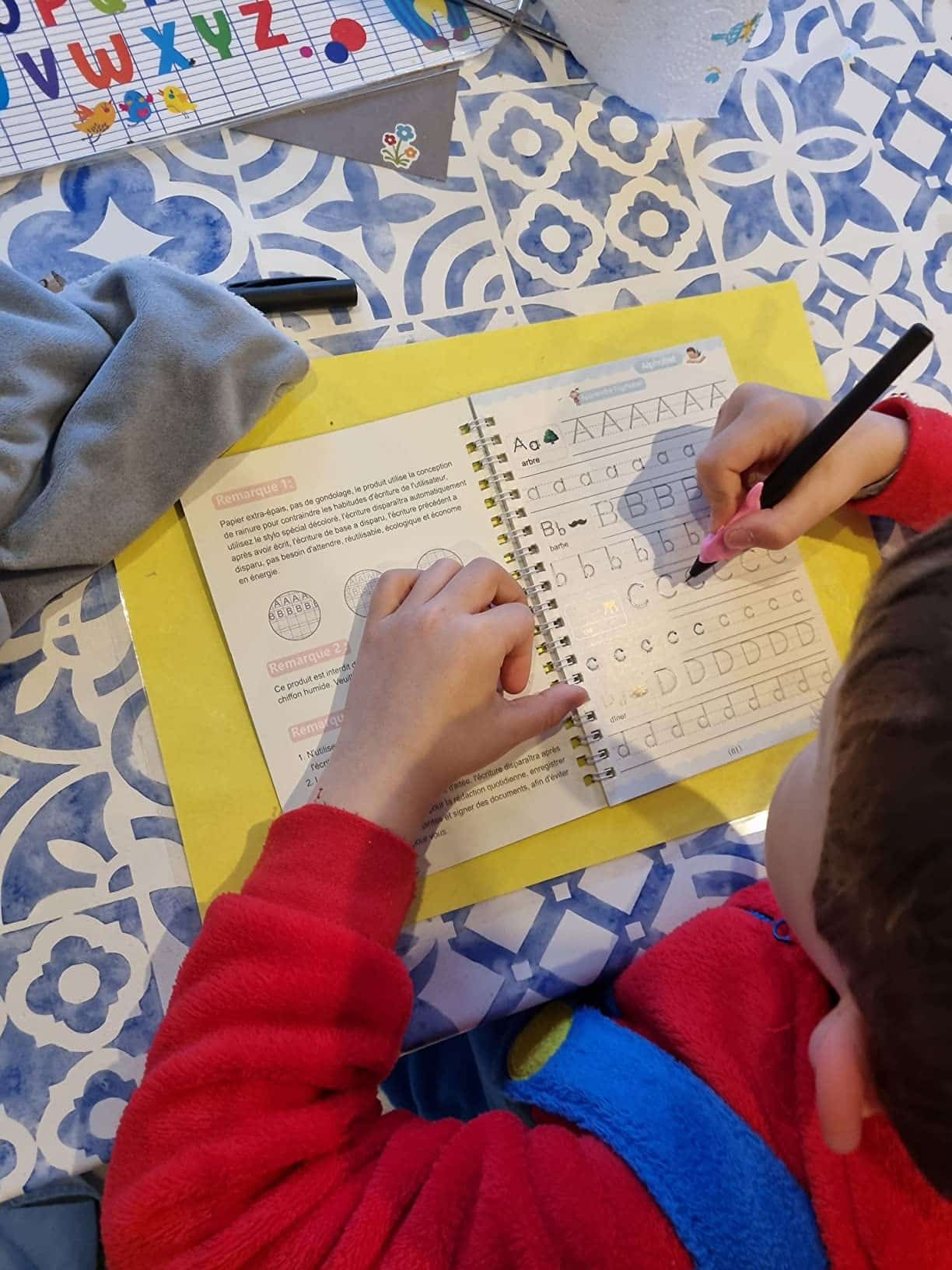 Un enfant s'exerce assidûment à l'écriture manuscrite dans un cahier d'exercices, traçant des lettres au crayon. La table, ornée d'une nappe à motifs bleu et blanc, accueille également un kit sensoriel coloré pour stimuler son apprentissage.