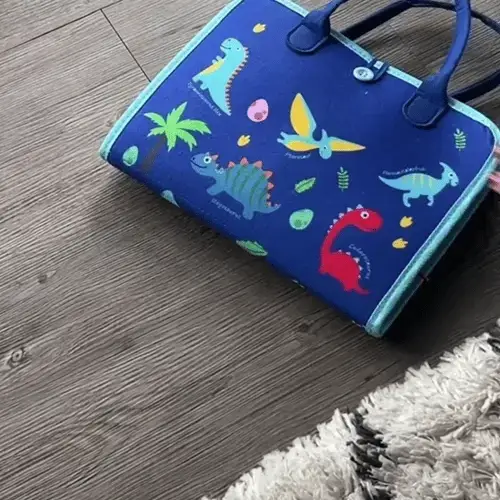 Sac bleu avec des illustrations colorées de dinosaures sur un plancher en bois à côté d'un tapis moelleux.