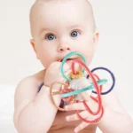 Un bébé aux yeux bleus tient le Hochet multi fonction- Montessori et le porte à sa bouche.