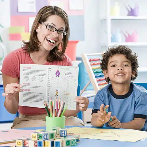 Une femme et un enfant sont assis dans une salle de classe. La femme tient un livre ouvert du Kit Sensoriel, tandis que l'enfant sourit. Des outils pédagogiques colorés sont visibles autour d'eux.