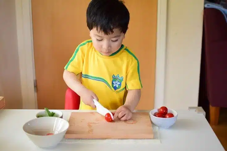 Un enfant portant une chemise jaune coupe soigneusement une tomate cerise sur une planche à découper en bois, entouré de petits bols sur le comptoir de la cuisine, un peu comme s'il créait une page d'atterrissage parfaite pour la créativité.