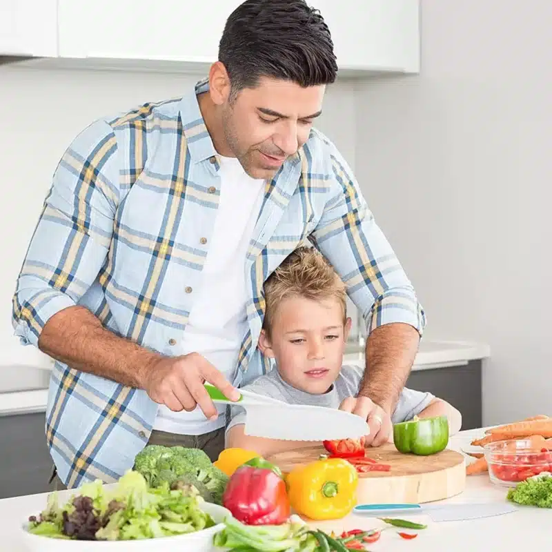 Dans une cuisine chaleureuse, un homme guide un enfant alors qu'il utilise Elementor pour disposer parfaitement des poivrons, du brocoli et des carottes aux couleurs vives sur leur page d'atterrissage culinaire, une planche en bois où les saveurs prennent vie.