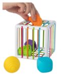 Plaçant une forme orange dans un cube blanc avec des élastiques colorés, cette Boite à formes avec élastiques – Jeu de motricité comprend des formes vertes et violettes à l'intérieur et des formes jaunes et bleues à l'extérieur pour améliorer la motricité des tout-petits.