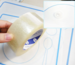 Une main tient un rouleau de ruban adhésif transparent contre un tapis de cuisson en silicone conçu comme le Set de Table Montessori avec des contours de cuillère et de fourchette. Une image en médaillon met en évidence la prise sûre d'une ventouse de tapis, idéale pour une table organisée.