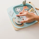 Une main navigue dans le « Puzzle pêche en bois magnétique », en utilisant une canne à pêche magnétique pour ramasser des morceaux d'étoiles de mer, de crabe et de poisson sur un fond clair sur le thème de l'océan.