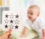 Une personne montre un « Jouet montessori imagier carte pour les bébé » comportant des étoiles noires devant un bébé flou vêtu de vêtements blancs, évoquant une scène inspirée de Montessori.