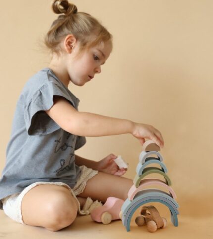 Une jeune fille avec un chignon empile des arches Arc en Ciel Montessori en Silicone aux couleurs pastel sur une surface claire. Elle est accompagnée d'une petite voiture et de blocs en silicone, vêtue d'un t-shirt gris et d'un short clair sur un fond neutre.