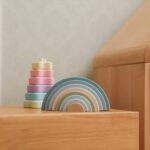 Une configuration Montessori enchanteresse comprend un empileur arc-en-ciel Montessori en silicone Arc en ciel et une tour à anneaux de couleur pastel sur une surface en bois, complétée par un fond mural clair.