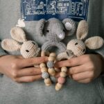 Une personne tient de charmants jouets animaux crochetés, dont un éléphant et deux lapins, avec des anneaux de dentition en perles de bois de « Anneaux de dentition Animaux – bois et crochet » contre un sweat-shirt gris, présentant un mélange enchanteur de bois et de crochet.