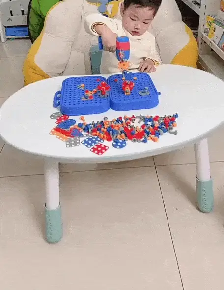Un jeune enfant est assis à une table et utilise une perceuse jouet pour assembler des pièces de construction en plastique colorées sur un tableau bleu.
