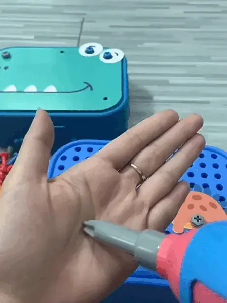 Une main est ouverte, un tournevis en plastique est pointé vers la paume. Des boîtes à jouets bleues aux visages de dessins animés apparaissent en arrière-plan.