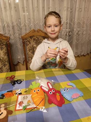 Un enfant est assis à une table avec du matériel de couture et des formes d'animaux en feutre coloré, évoquant une charmante boîte à outils. En arrière-plan, des rideaux moelleux et des chaises encadrent cette scène de bricolage chaleureuse.