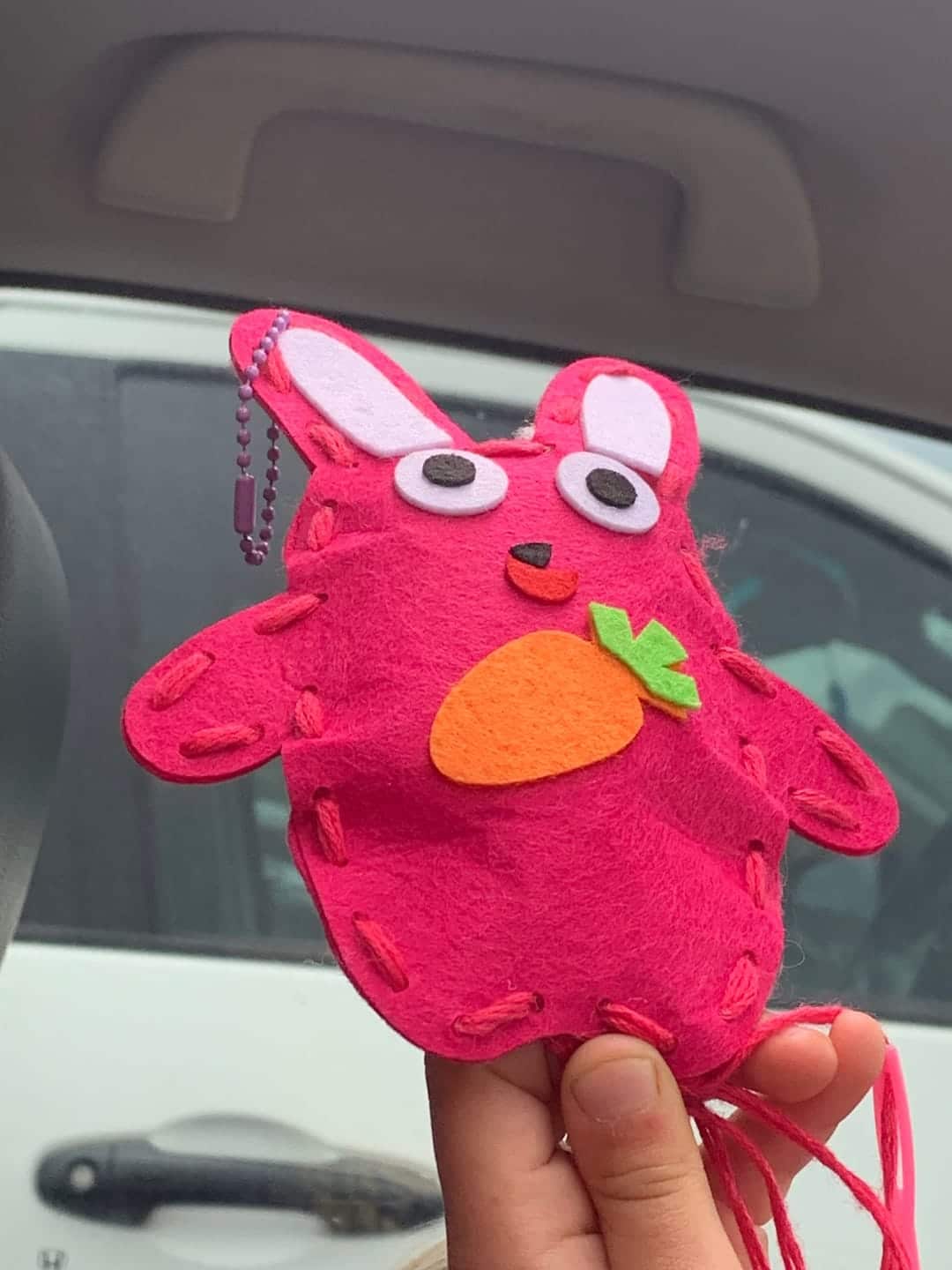 Une main tient un lapin en feutre rose fait main, décoré d'une carotte, avec des yeux noirs et une chaîne, suspendu comme un charme inattendu au milieu d'outils utilitaires. L'arrière-plan révèle l'intérieur d'une voiture et une partie d'un véhicule blanc.
