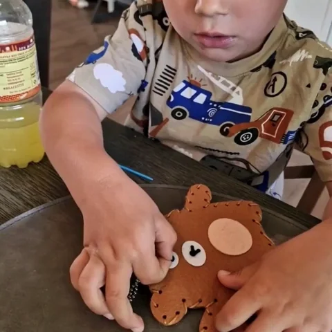 Un enfant utilise joyeusement des outils pour assembler un objet en forme d'ours sur la table, avec une bouteille de boisson jaune vif à proximité.
