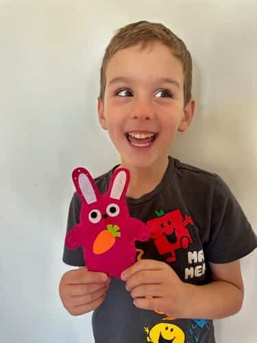 Un enfant, tout sourire, tient un objet en feutre rose en forme de lapin avec une carotte, tout en exhibant fièrement sa nouvelle boîte à outils. Il porte un t-shirt foncé aux motifs colorés, parfait pour ses premières aventures de bricolage.
