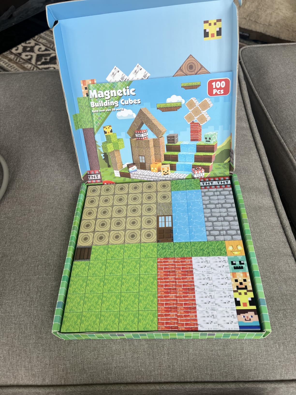 Une boîte de « cubes de construction magnétiques » contenant 100 pièces est ouverte, montrant des tuiles à motifs de style Minecraft disposées soigneusement à l'intérieur.