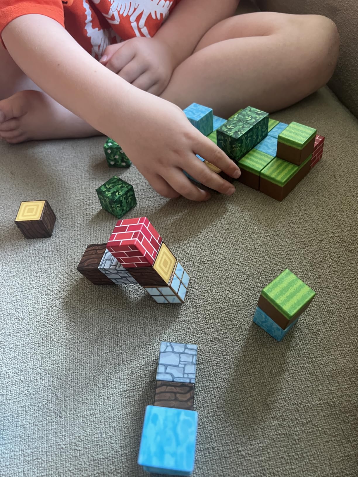 Un enfant est assis sur un canapé, jouant avec des blocs de construction sur le thème de Minecraft, les disposant d'une main tandis que d'autres blocs sont dispersés à proximité.