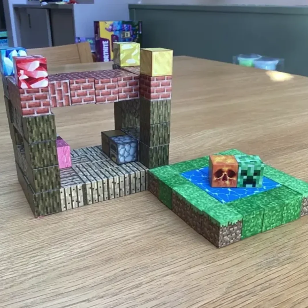 Deux modèles en papier sur le thème de Minecraft sont posés sur une table en bois : une structure semblable à une maison et une petite piscine avec des blocs Creeper et citrouille.